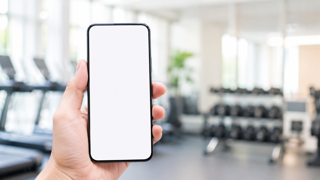 明るいジムで、画面に何も表示されていないスマートフォンを片手で持っている様子